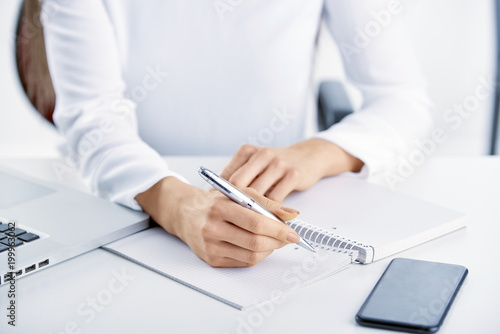 Closeup photo of businesswoman s hand holding pen and writing something while using mobile phone