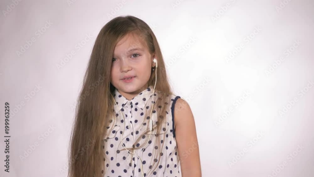 Child with earflaps dancing at studio background. The girl listens to music on the smartphone. The kid has loose long hair. Shooted on a gray white background. Child with earflaps dancing at studio background. The girl listens to music on the smartphone. The kid has loose long hair. Shooted on a gray white background.