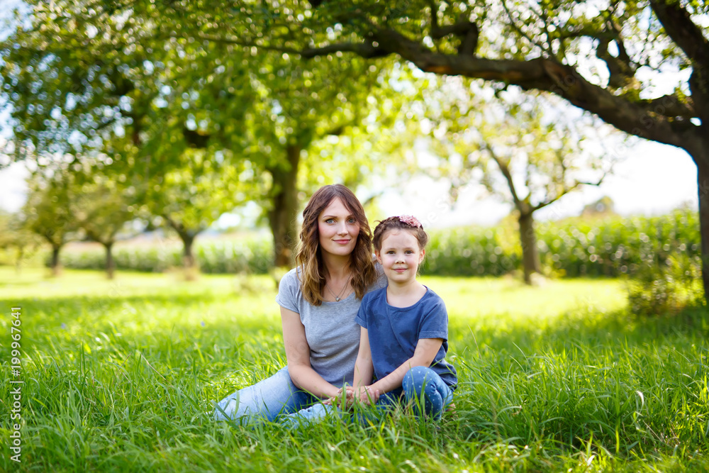 Beautiful young mother and little daughter sitting on green grass and resting Beautiful young mother and little daughter sitting on green grass and resting