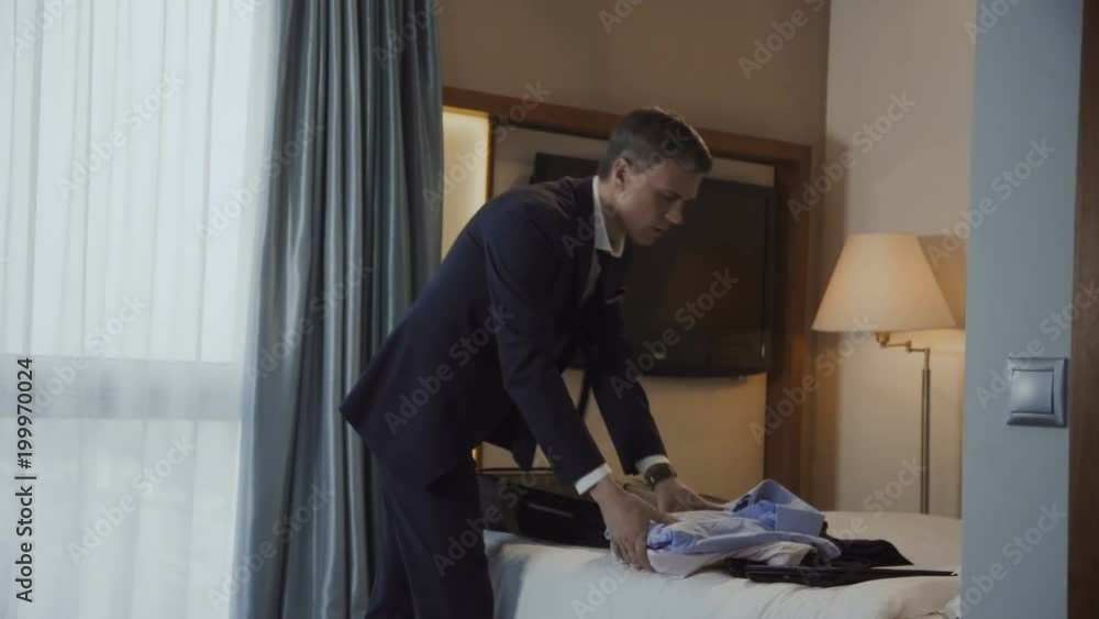 Adult man in suit having phone call meanwhile packing suitcase leaving hotel room at end of business trip. Adult man in suit having phone call meanwhile packing suitcase leaving hotel room at end of business trip.