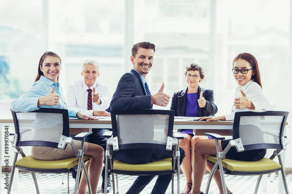 Successful business team showing thumbs up and looking at camera. Cheerful colleagues working as team. They sitting at table and showing approval sign. Meeting concept