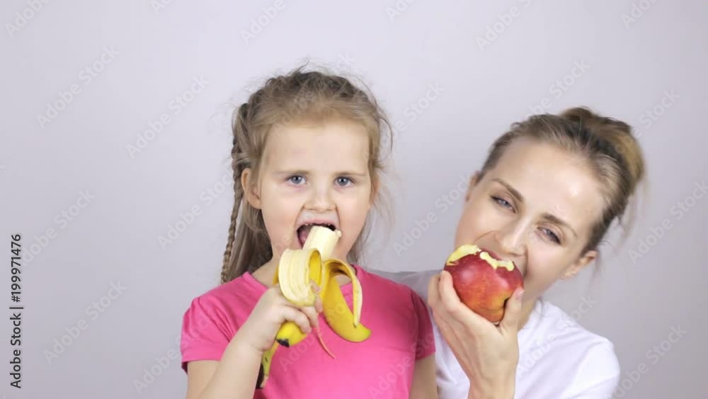 Young mother with her daughter smiling and eating banana and apple Young mother with her daughter smiling and eating banana and apple