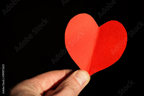 Red heart made from paper in the hand of a young man on a black background.