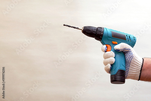hand in dotted work glove holding cordless drill machine on gray background, copy space