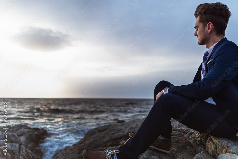 Pensive man in suit sits on the rocks near sea and thinking. Young guy meditating on background of the seascape and skytest