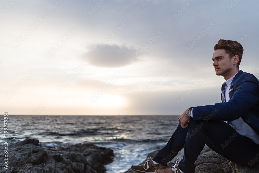 Serious young man in elegant suit sits on the coast near ocean. Young guy thinking on background of the water and sky