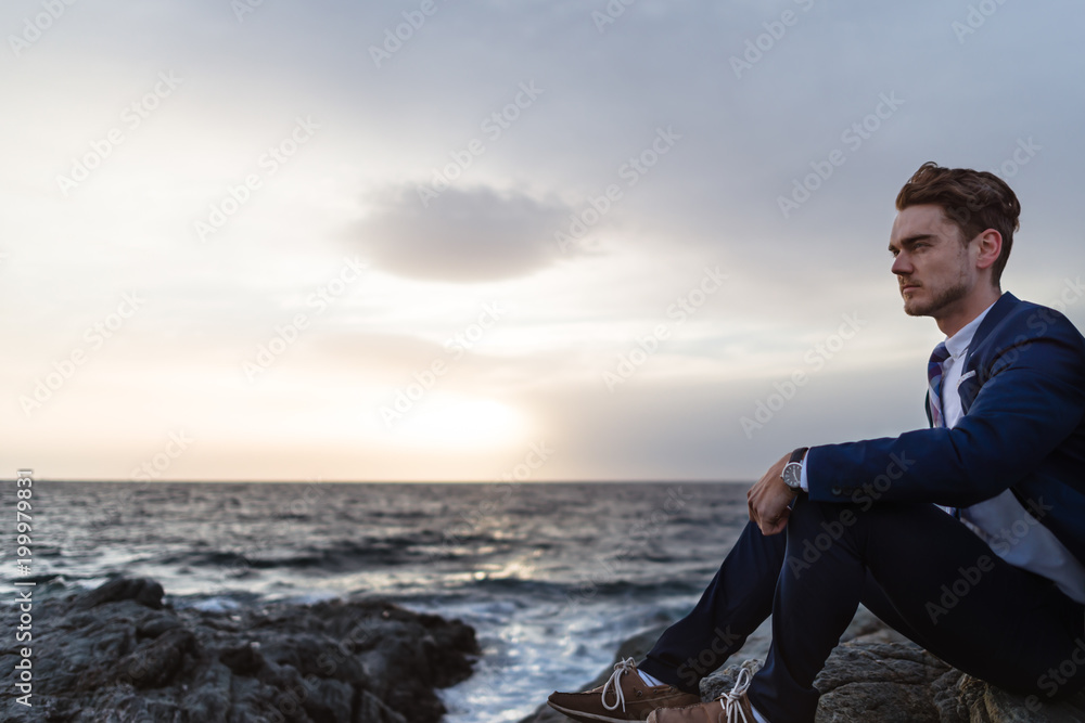 Serious young man in elegant suit sits on the coast near ocean. Young guy thinking on background of the water and sky Serious young man in elegant suit sits on the coast near ocean. Young guy thinking on background of the water and sky