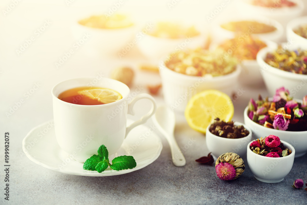 Tea concept with copy space. Different kinds of dry tea in white ceramic bowls and cup of aromatic tea on grey background. Tea concept with copy space. Different kinds of dry tea in white ceramic bowls and cup of aromatic tea on grey background.