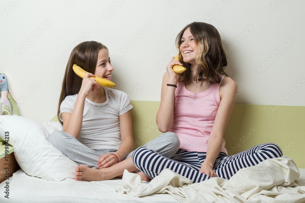 Two sister children in pajamas play in the morning in bed. Keep bananas as phones talking and laughing.