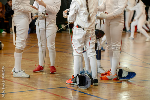 Girls, participants in fencing competitions on swords stand in the center of fencing hall waiting for beginning of the fight. Fencing masks on the floor, swords in their hands.
