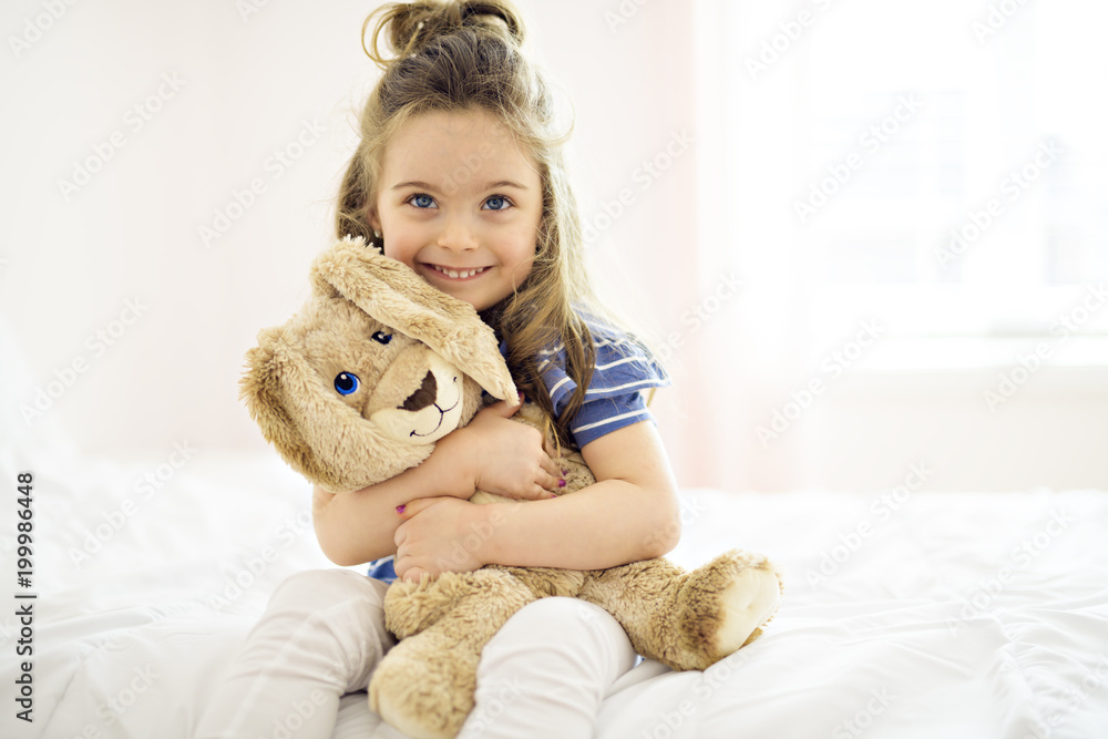 Sweet little girl is hugging a teddy bear on her bed at home