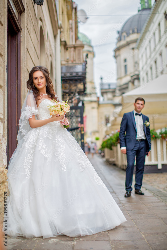 Beautiful bride in white wedding dress is holding bouquet of roses, behind her is groom in tuxedo. Romantic walk around the city