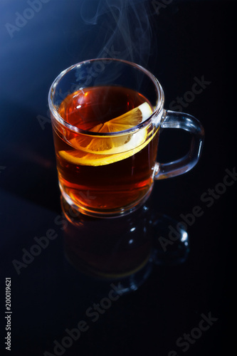A glass mug of tea with lemon stands on a black glass. The tea is hot, steam is coming. A cup of tea is beautifully reflected in the glass
