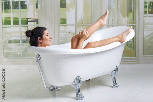 Happy laughing asian woman shaving her legs and having fun while lying in bathtub