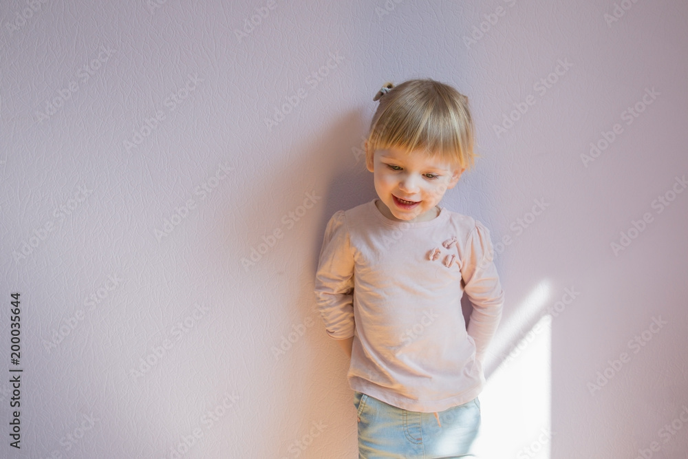 Happy little blond girl stands leaning against the wall in a sunny room at home. She looks at the viewer and laughs joyfully. Close up shot. Copy space.