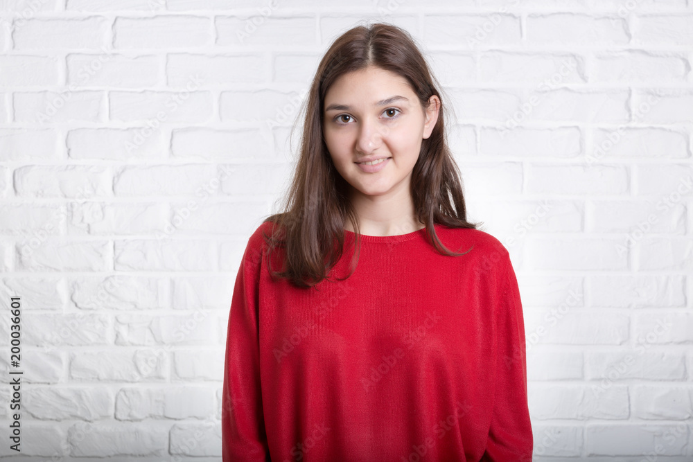 A young pretty smiling girl in a red sweater and a hat against a modern white brick wall in the loft style. She looks at the viewer. Copy space. A young pretty smiling girl in a red sweater and a hat against a modern white brick wall in the loft style. She looks at the viewer. Copy space.