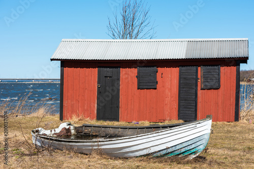 White murky rowboat on the ground in front of a red and black wooden shed in coastal landscape. location Farjestaden on Oland, Sweden. photo