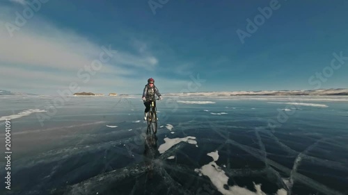 Woman is riding bicycle on the ice. The girl is dressed in a silvery down jacket, cycling backpack and helmet. Ice of the frozen Lake Baikal. The tires on the bicycle are covered with special spikes photo