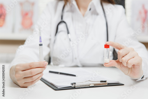 Female doctor sitting at desk with medical documents holds syringe with needle, bottle with medicine in light office in hospital. Woman in medical gown in consulting room. Healthcare, medicine concept