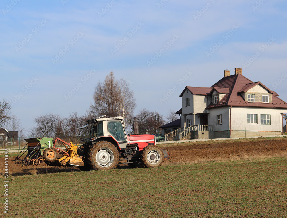 Fototapeta premium A farmer on a red tractor with a seeder sows grain in plowed land in a private field in the village area. Mechanization of spring field work. Farmer's everyday life. Processing of land. The agrarian.