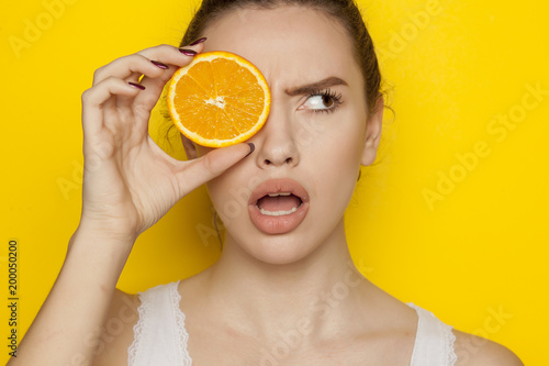 Surprised young woman posing with slice of orange on her face on yellow background