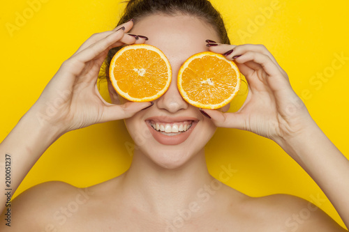 Happy young woman posing with slices of oranges on her face on yellow background