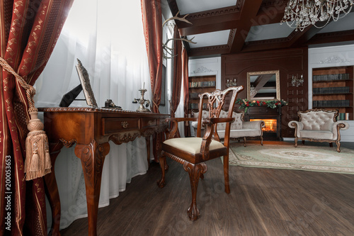 Old studying room with fabric armchairs and wooden table. Classic bookcase filled with books. Fireplace