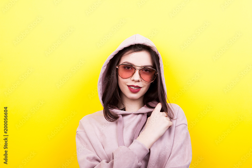 Cool caucasian woman wearing a hood and eyeglasses on yellow background