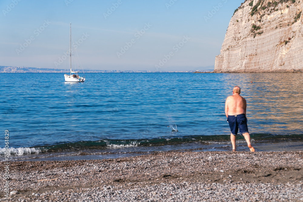 an elderly man throws stones at sea, on the beach