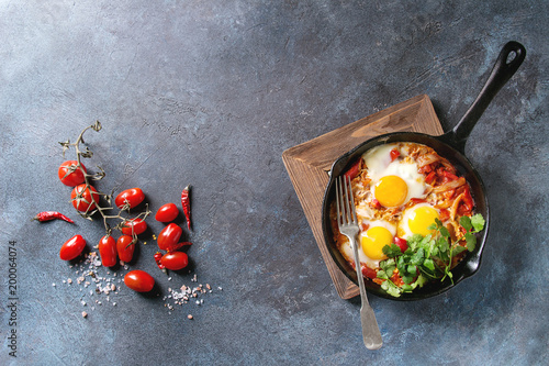 Traditional Israeli Cuisine dishes Shakshuka. Fried egg with vegetables tomatoes and paprika in cast-iron pan on wooden board with coriander herbs over blue texture background. Top view, space.
