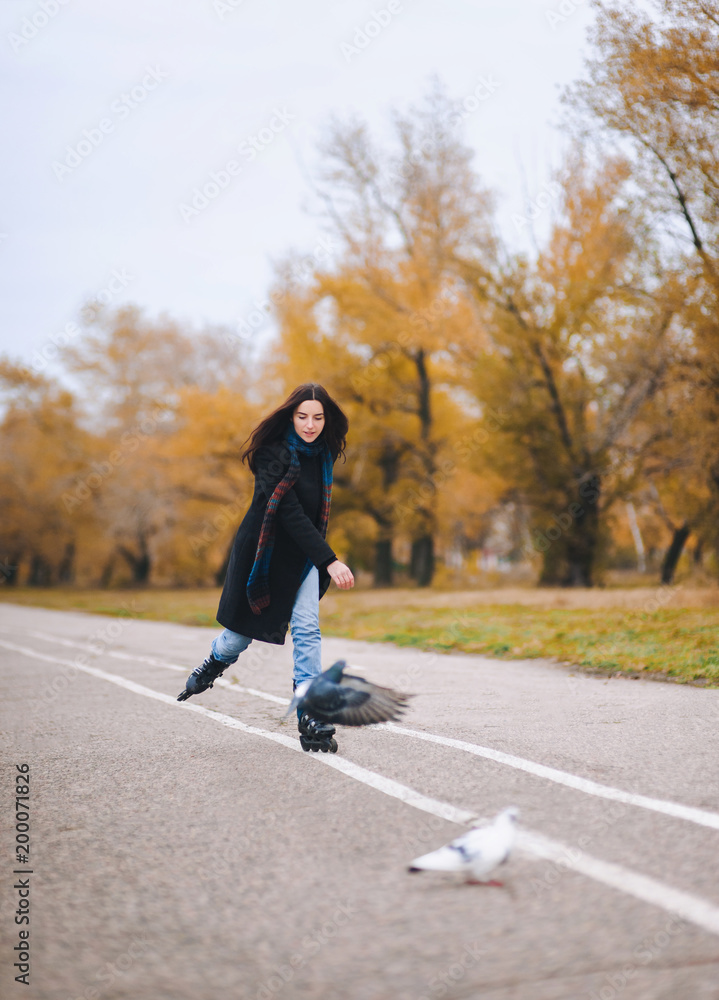 A pretty young girl in a coat and blue jeans with a long scarf happily skates in the autumn park with pigeons. Fighting the autumn depression. A pretty young girl in a coat and blue jeans with a long scarf happily skates in the autumn park with pigeons. Fighting the autumn depression.