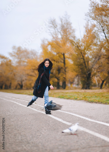A pretty young girl in a coat and blue jeans with a long scarf happily skates in the autumn park with pigeons. Fighting the autumn depression.