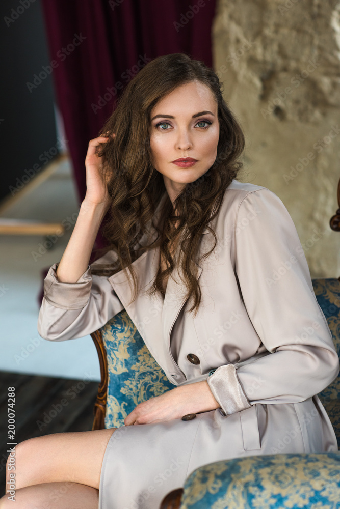 elegant woman in fashionable trench coat sitting in armchair