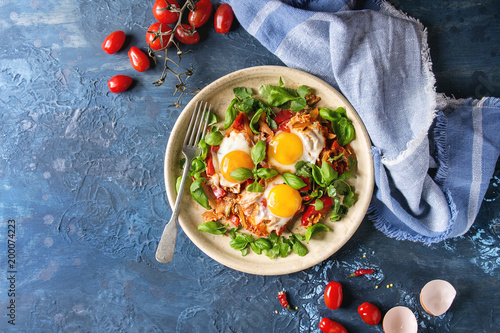 Traditional Israeli Cuisine dishes Shakshuka. Fried egg with vegetables tomatoes and paprika in ceramic plate with cloth, herbs and ingredients above over blue texture background. Top view, space.