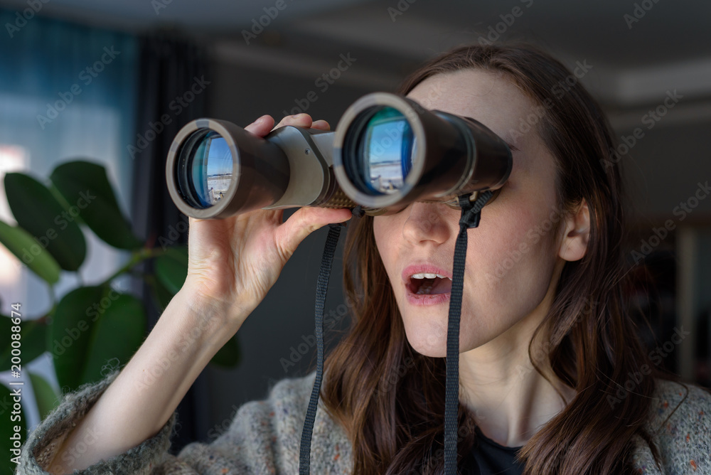 Portrait of a surprised brunette with binoculars looking out the window, spying on neighbors Portrait of a surprised brunette with binoculars looking out the window, spying on neighbors