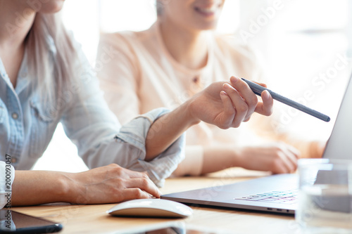 Close up shot of two female businesswomen working with hands on a laptop keyboard in an office in sunlight at a wooden table and one of them showing a pen on the display