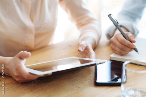 Two female businesswomen working with hands on a tablet in an office in sunlight at a wooden table and one of them writing a pen in a stylish blank on the desk is a modern smartphone