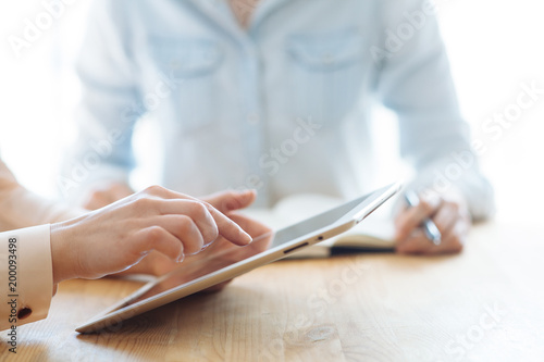 Two female businesswomen working their hands on a tablet in an office in the sunlight at a wooden table and one of them writes a pen in a stylish notebook, and touches the tablet with her finger