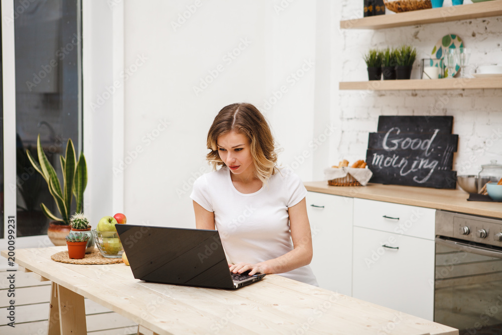 A beautiful girl is sitting at the table in the kitchen and looks at the laptop computer monitor, typing text. Modern technological devices. the Internet A beautiful girl is sitting at the table in the kitchen and looks at the laptop computer monitor, typing text. Modern technological devices. the Internet