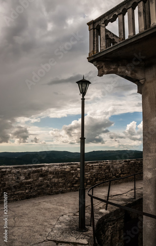 An empty old mediterranean street with cracks on the floor, a lamp post, a fragment of a balcony under a cloudy gray sky. The photo was shot in Motovun town, Croatia.
