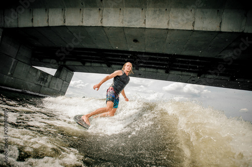 Athletic long-haired man wakesurfing on the board under the concrete bridge