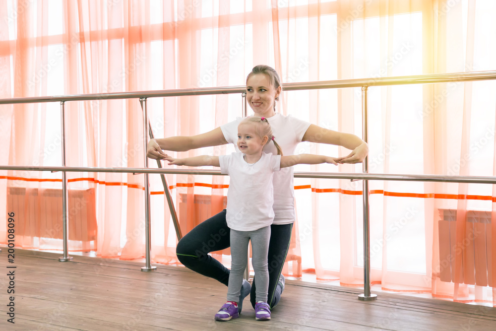Fototapeta premium A little girl and her mother are doing sports. A young woman with a daughter doing a sports exercise - stretched her arms out to the sides.