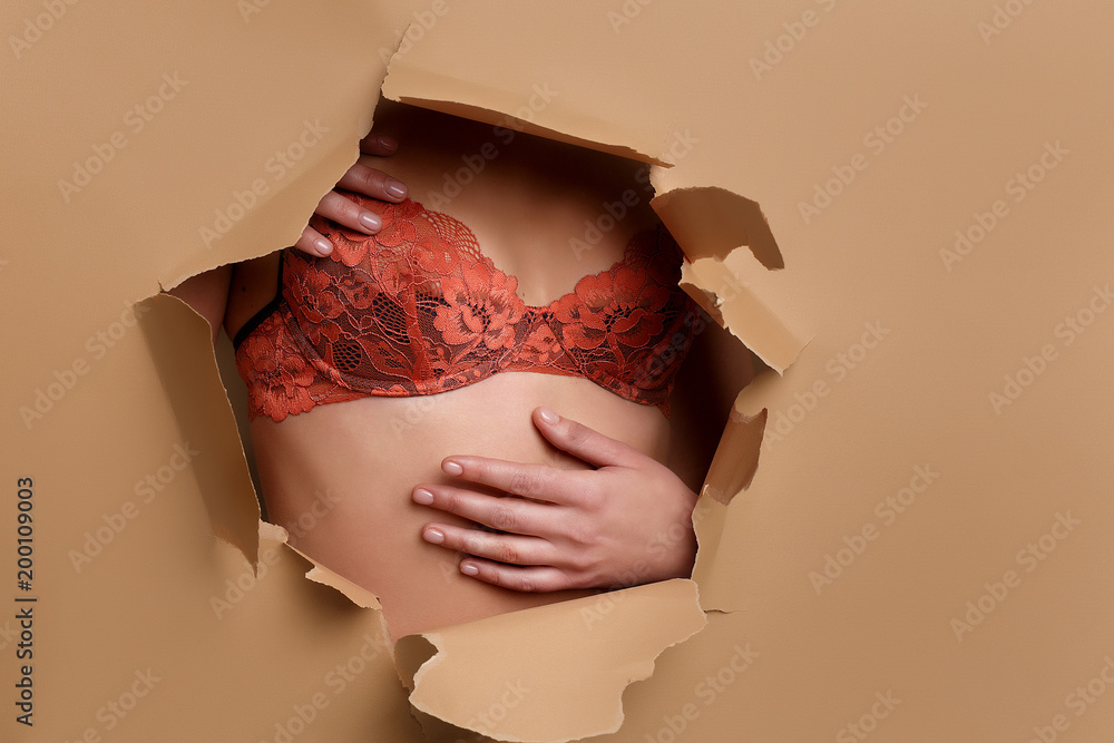 close-up of a girl's bust in an orange lace bra that looks through a broken  hole in beige paper Stock Photo