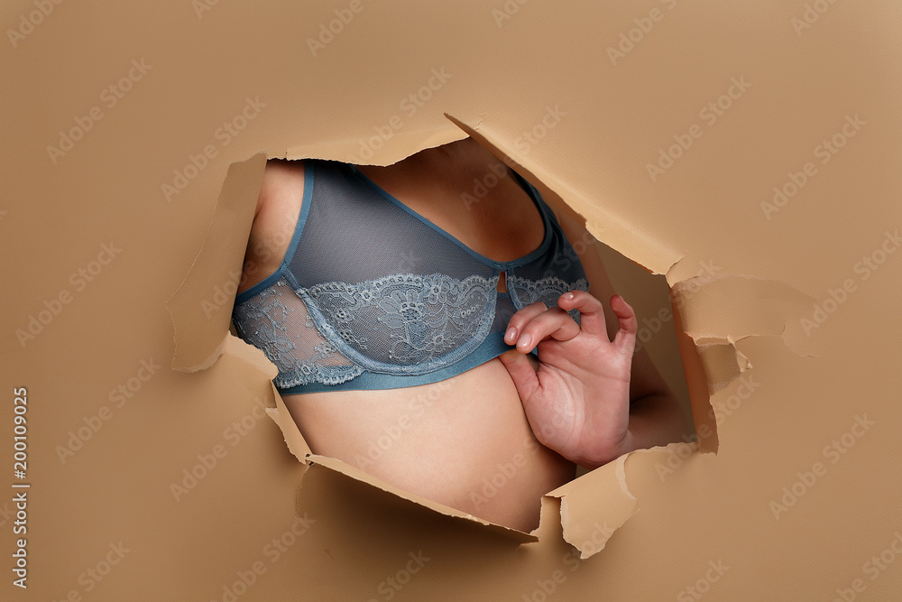 close-up of a girl's bust in a blue lace bra that looks through a broken  hole in beige paper Stock Photo