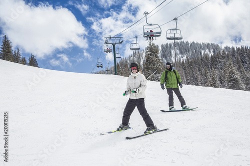 A young skier and a snowboarder skiing and boarding down a ski slope with the chair lift in the background. A fun winter day at the ski resort