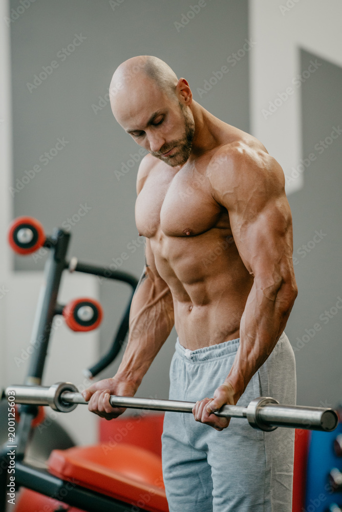 Fototapeta premium Shredded shirtless bodybuilder with beard and shaved head doing biceps curls with barbell in a gym. Fit man trains.