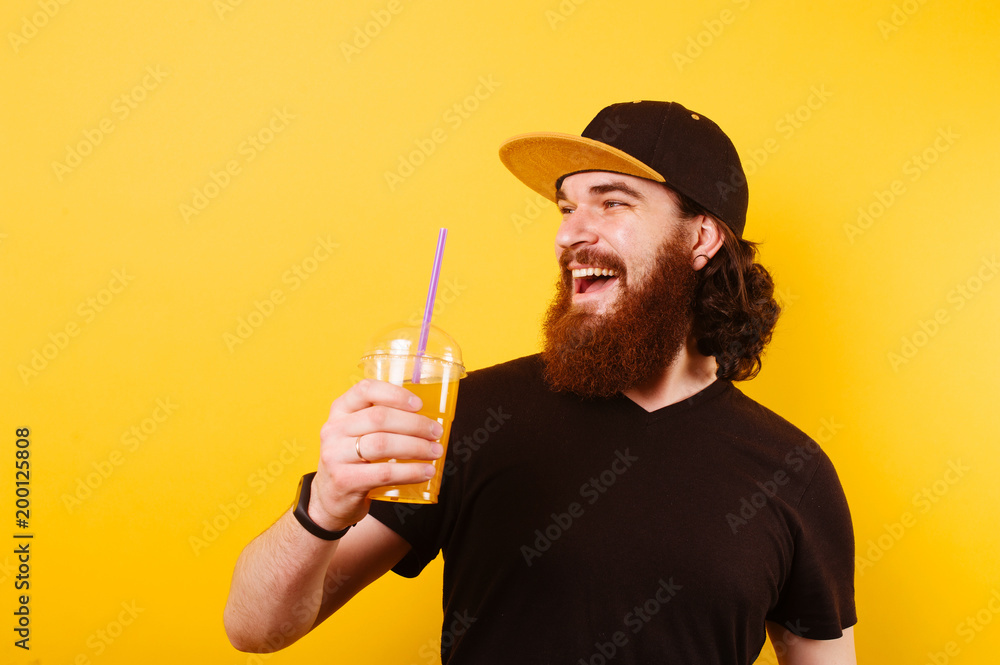 Cheerful bearded hispter man with cap holding plastic cup of fresh orange  juice and looking away over yellow background Stock Photo | Adobe Stock