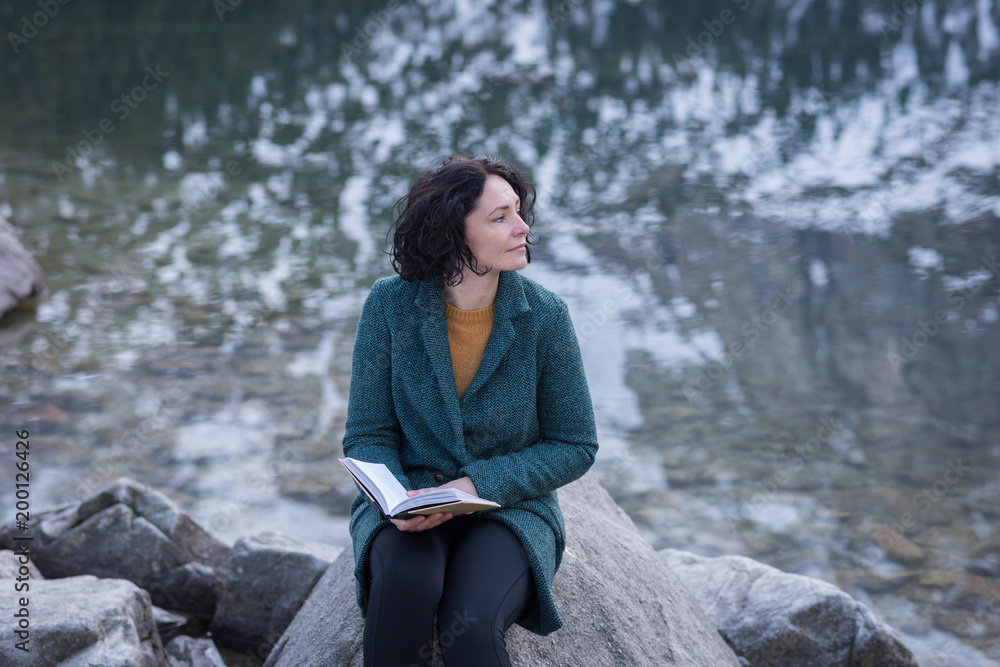 Thoughtful woman sitting with book at lakeside Thoughtful woman sitting with book at lakeside