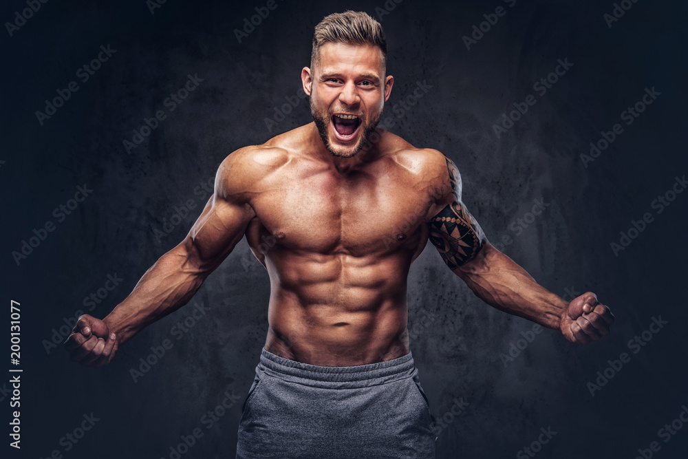 A handsome shirtless tattooed bodybuilder with stylish haircut and beard, wearing sports shorts, posing in a studio. Isolated on a dark background