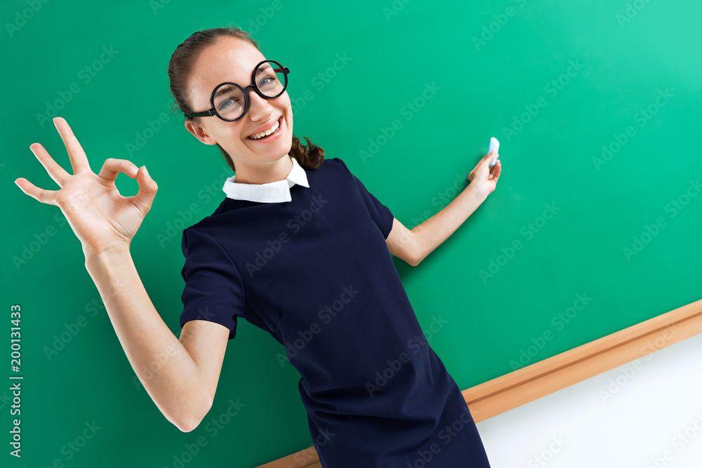 Young teacher shows gesture okay and points to something. Photo of attractive girl near blackboard, education concept